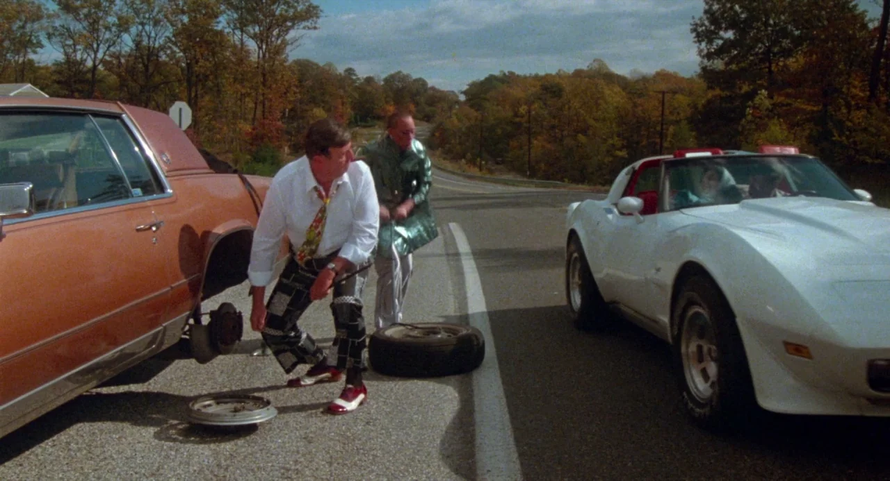 Polyester (1981) movie still: wide — A rural asphalt road lined with trees during an autumn afternoon. Two men are attending to a car with a flat…