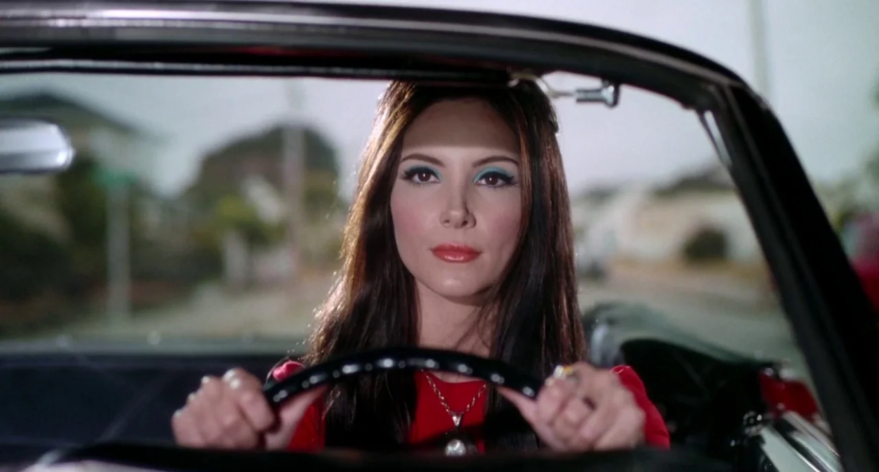 The Love Witch (2016) movie still: medium — A woman sits inside a car on a bright daytime road. She grips the black steering wheel with both hands…