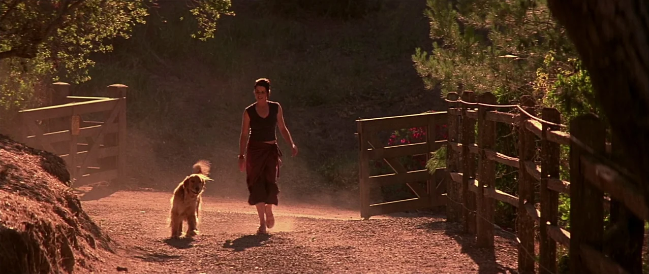 Scream 3 (2000) movie still: fullbody — A gravel path leads through a rural area with wooden fences on both sides. A woman walks toward the camera…
