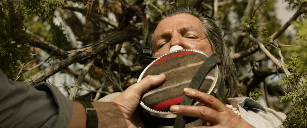 The Marksman (2021) movie still: closeup — A man with long grey hair lies beneath dense foliage in a forest setting. An unseen second person holds a…