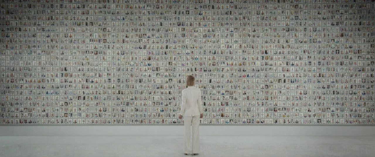 I Care A Lot (2020) movie still: wide — A spacious gallery features a wall covered in hundreds of small, grid-aligned portrait photographs. A woman…