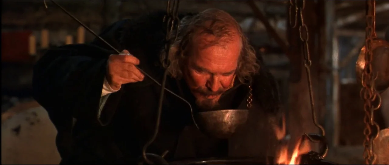 Ravenous (1999) movie still: medium — A dimly lit medieval hearth with chains and cooking equipment hanging above a fire. An older man in a dark…