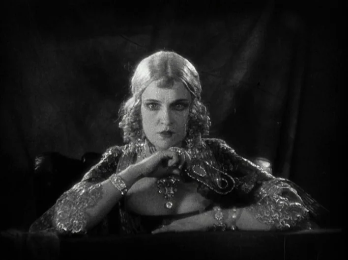 The Man Who Laughs (1928) movie still: medium — A woman sits at a dark table draped with a plain, heavy fabric background. She stares forward with…