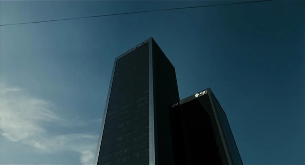 Sidewalls (2011) movie still: establishing — Two modern glass-clad office towers reach upward against a clear blue sky. A single power line cuts…