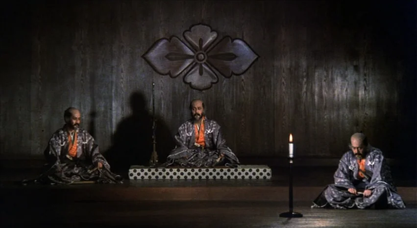 Kagemusha (1980) movie still: wide — Three men in traditional robes sit in a row on floor mats. A dark wooden wall features a large, symmetrical…