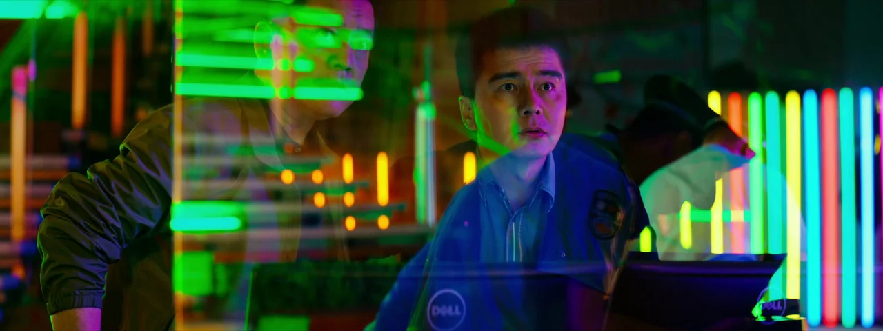 Under The Light (2023) movie still: medium — A dark room illuminated by rows of vibrant vertical neon lights and glowing screens. Two men monitor…