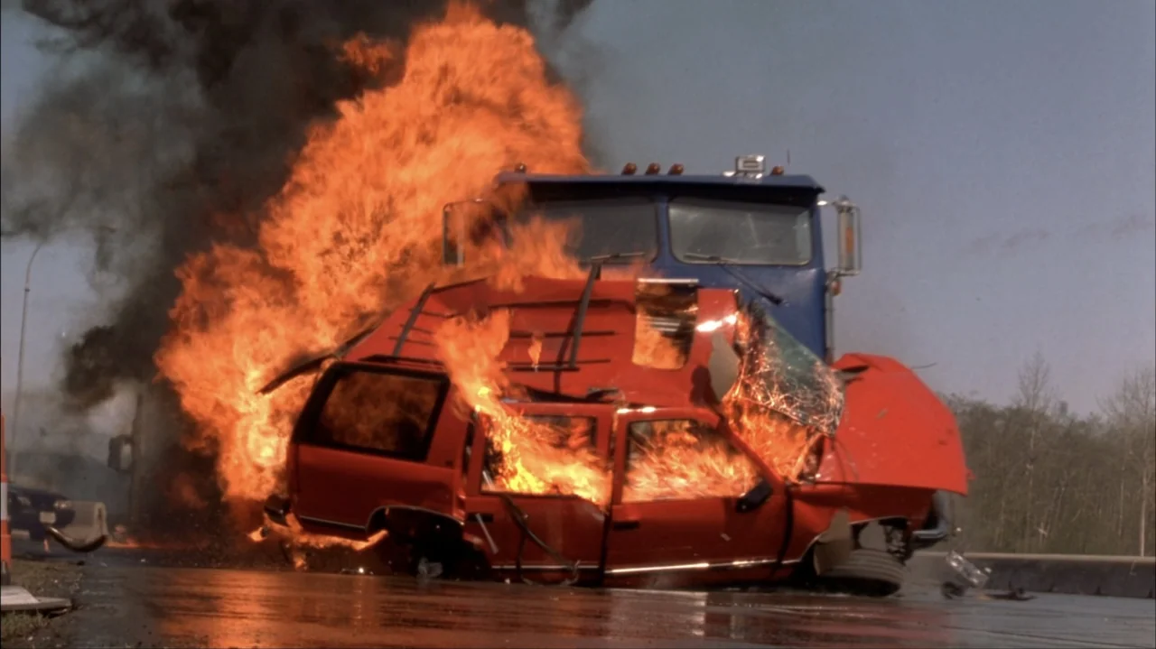 Final Destination 2 (2003) movie still: medium — A mangled red SUV is engulfed in flames on an asphalt road surface. The burning wreckage sits in…