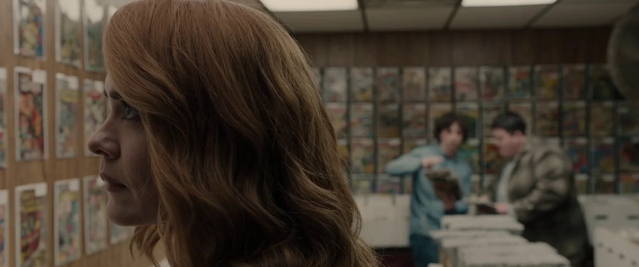 Glass (2019) movie still: closeup — A cluttered comic book store features shelves filled with bagged and boarded vintage comics. A woman in the…