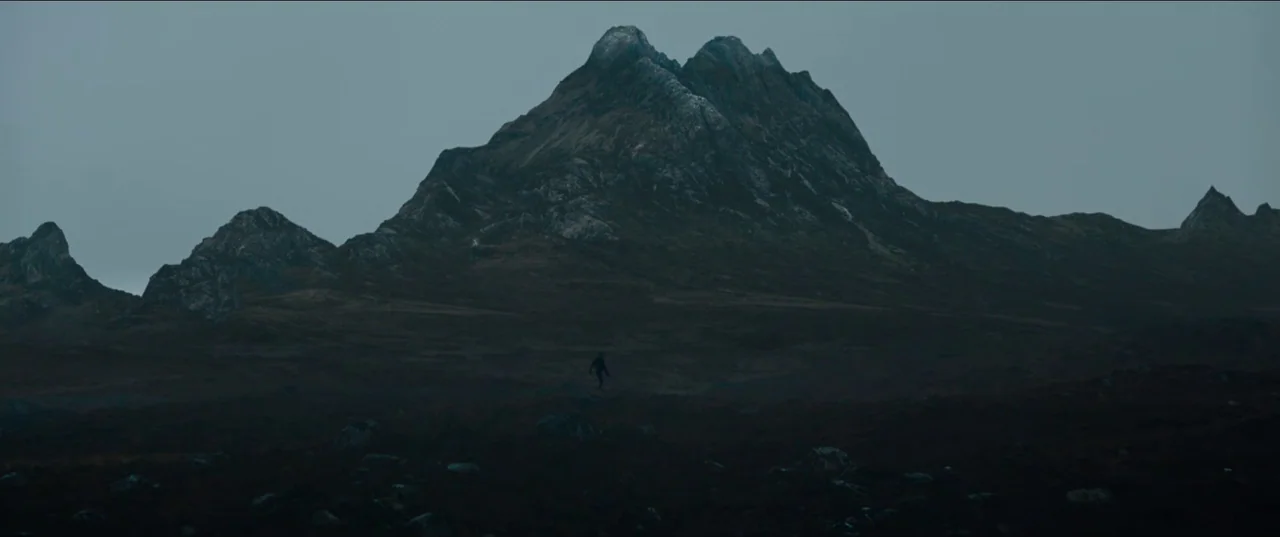 Out of Darkness (2022) movie still: wide — A vast, desolate mountain range spans the frame under a heavy, overcast sky. A lone figure walks across…