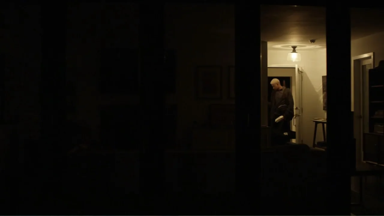Like.Share.Follow. (2017) movie still: medium — A dimly lit room is viewed through dark window frames from the outside. A man with light hair stands…