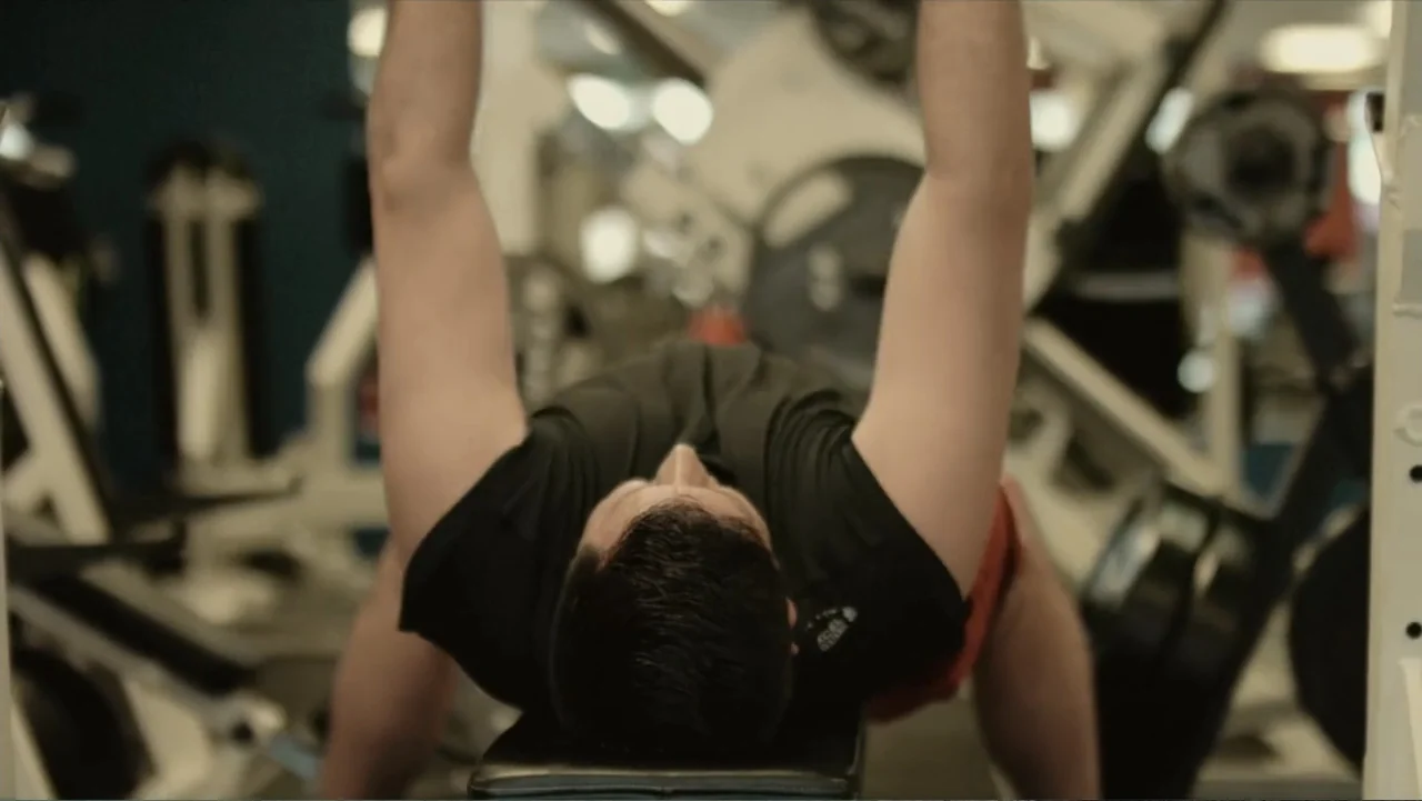 Death of a Child (2017) movie still: medium — A well-lit indoor fitness center featuring weight training equipment in the background. A man lies on…