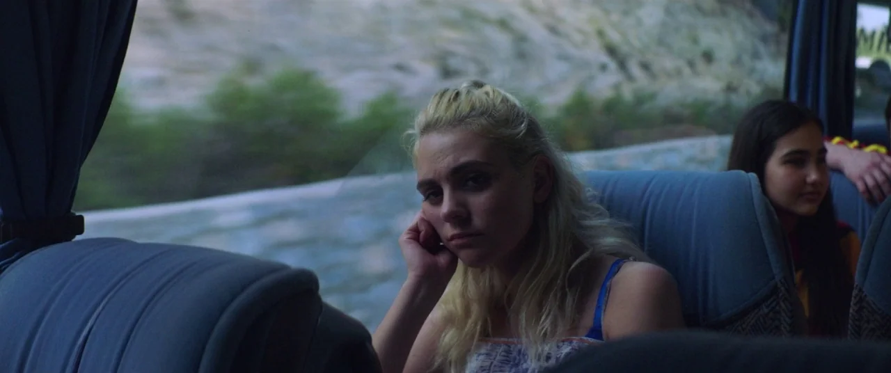 Holiday movie still: medium — The interior of a passenger bus with blue fabric seats and a window view. Two young women occupy the seats, one in the…