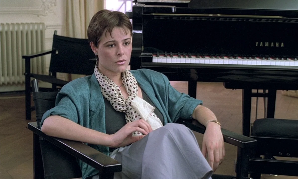 A Tale of Springtime (1990) movie still: medium — A well-lit room featuring a black Yamaha grand piano and director chair seating. A woman with…