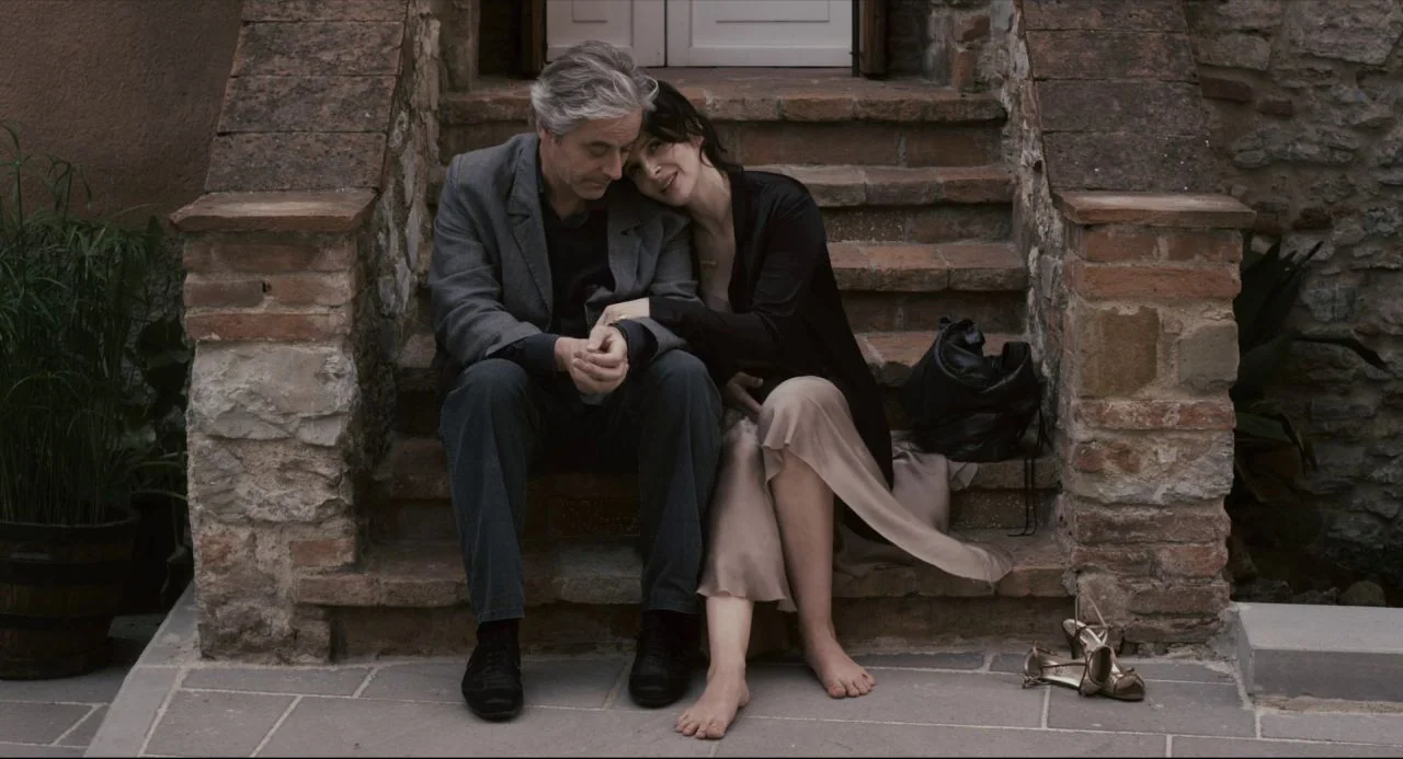 Certified Copy (2010) movie still: fullbody — A man and woman sit together on old stone steps beside a brick wall. They sit closely on the stairs…