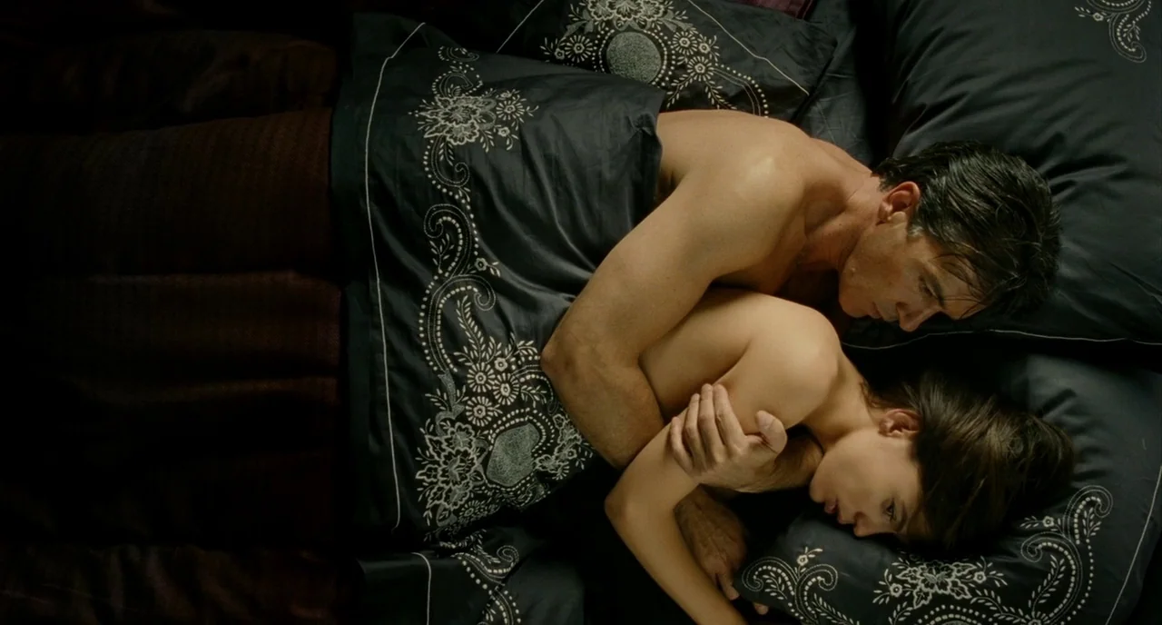 The Skin I Live In (2011) movie still: medium — A man and woman lie together in bed, covered by a black patterned quilt. The couple is embraced…