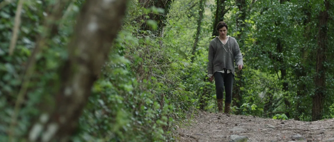 The Little Hours (2017) movie still: fullbody — A dense forest path winds through thick green vegetation with a prominent tree trunk. A young man…