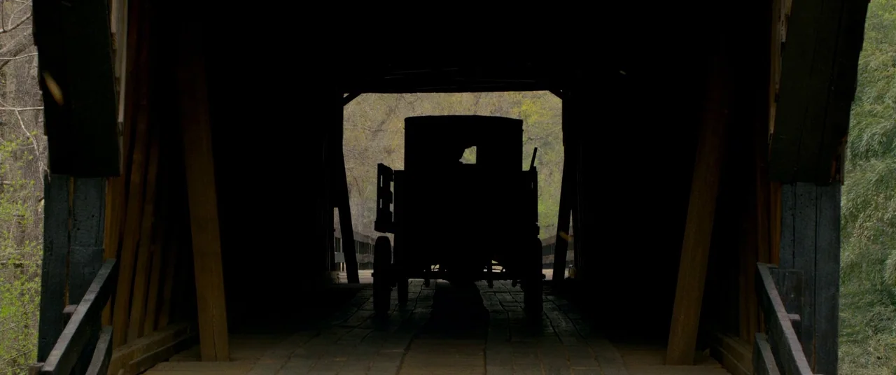 Lawless (2012) movie still: wide — A wooden covered bridge frames a dark carriage silhouetted against the bright exterior scenery. The horse-drawn…