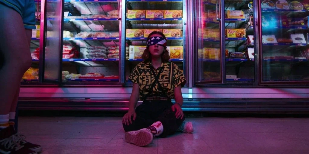 Stranger Things (2019) movie still: medium — A girl sits on the floor of a convenience store between large freezer aisles. She wears a blindfold…