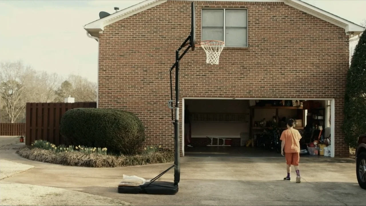 Death of a Child (2017) movie still: wide — A suburban residential driveway fronts a brick house with an open garage. A young boy in orange clothing…