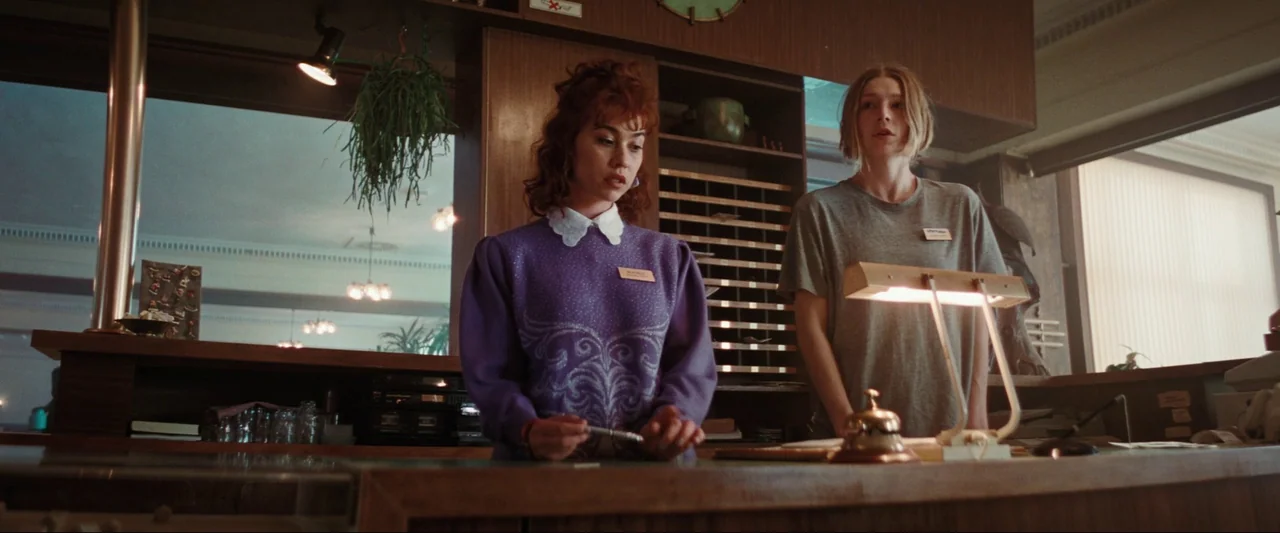 Cuckoo (2024) movie still: medium — A wood-paneled hotel reception counter displays cubbies, a desk lamp, and a metal bell. Two young women stand…