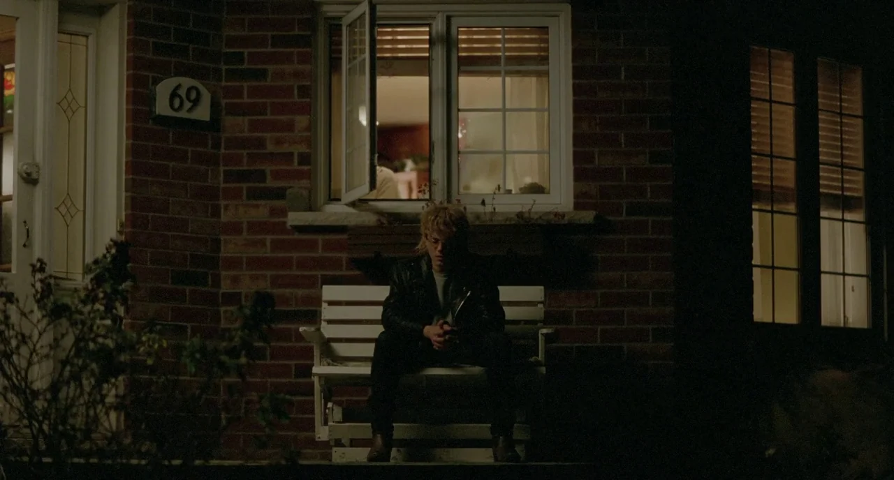 Tom at the Farm (2013) movie still: fullbody — A brick house exterior at night features a white wooden bench on the porch. A young person with blond…
