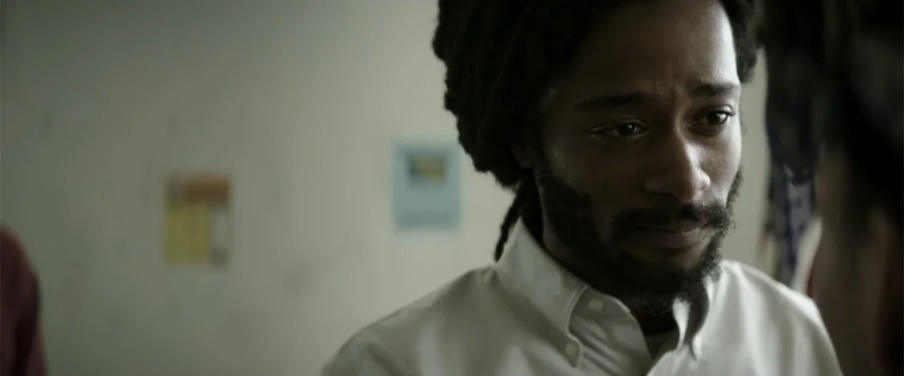 Crown Heights (2017) movie still: closeup — A spartan office interior features a white wall with a small framed notice. A Black man in a white…