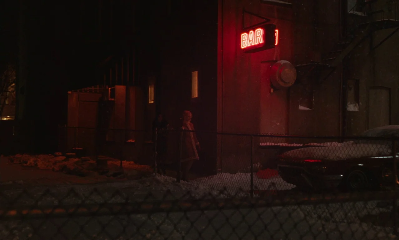 Eileen (2023) movie still: wide — A snow-covered alleyway features a dark wall with a bright red neon BAR sign. Two people stand behind a chain-link…