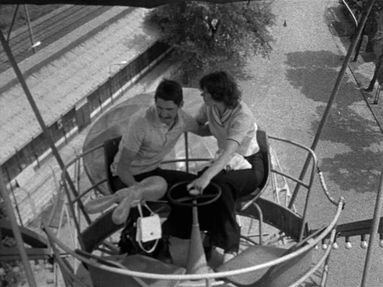 Family Nest (1979) movie still: medium — Two people sit inside a metal gondola ride high above a paved ground area. The couple faces each other…