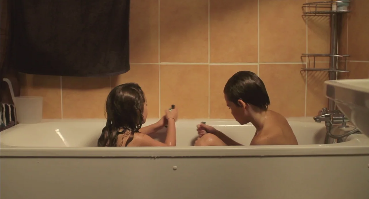Tomboy (2011) movie still: medium — A tiled bathroom with a white tub and a metal corner shelf unit. Two children are seated facing each other while…