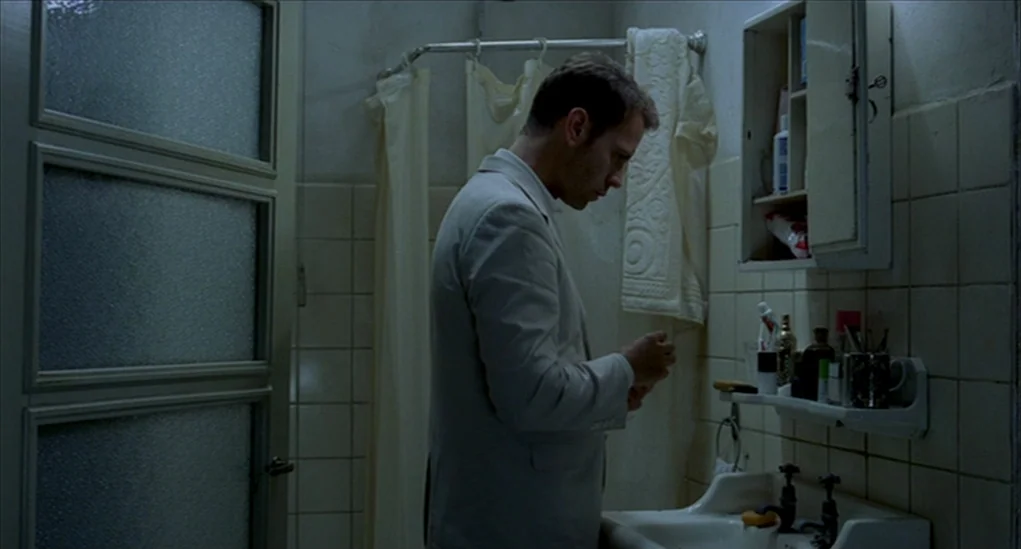 Anatomy of Hell (2004) movie still: medium — A dimly lit bathroom features tiled walls and a small medicine cabinet above a sink. A man in a blazer…