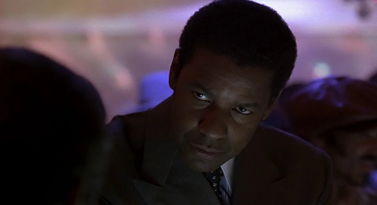 American Gangster (2007) movie still: closeup — A dimly lit nightclub with blurred lights in the background. A man in a suit looks toward another…