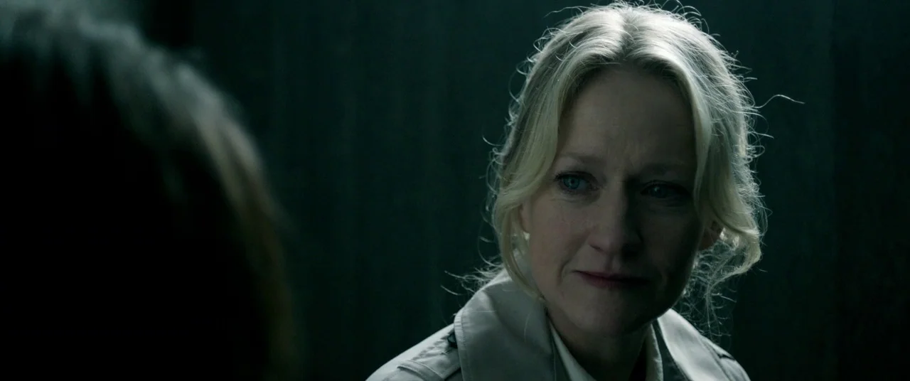 The Cured (2017) movie still: closeup — A dark, sparsely lit room with shadows obscuring the background. A blonde woman in a trench coat turns her…