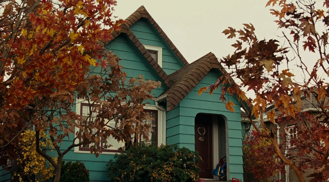 Juno (2007) movie still: establishing — A teal-sided house stands behind trees with vibrant yellow and orange autumn foliage. The structure features…
