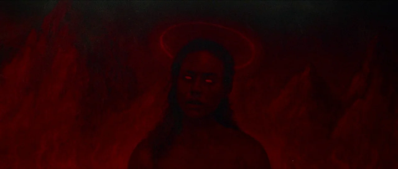 Bliss movie still: medium — A dark rocky landscape appears behind a woman against a deep red atmosphere. The woman faces forward with glowing eyes…