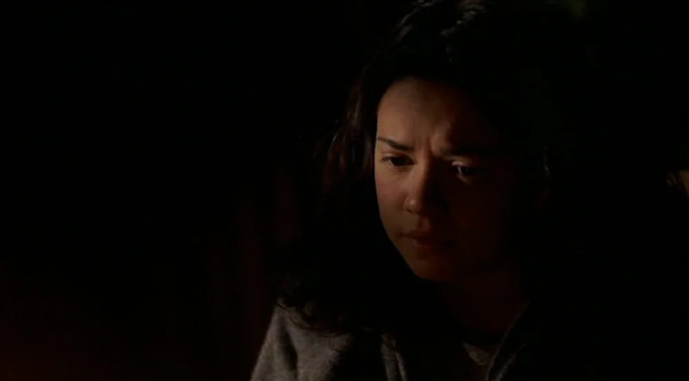 Limbo (1999) movie still: closeup — A dark, minimally lit interior space with shadows obscuring the surroundings. A woman with dark wavy hair looks…