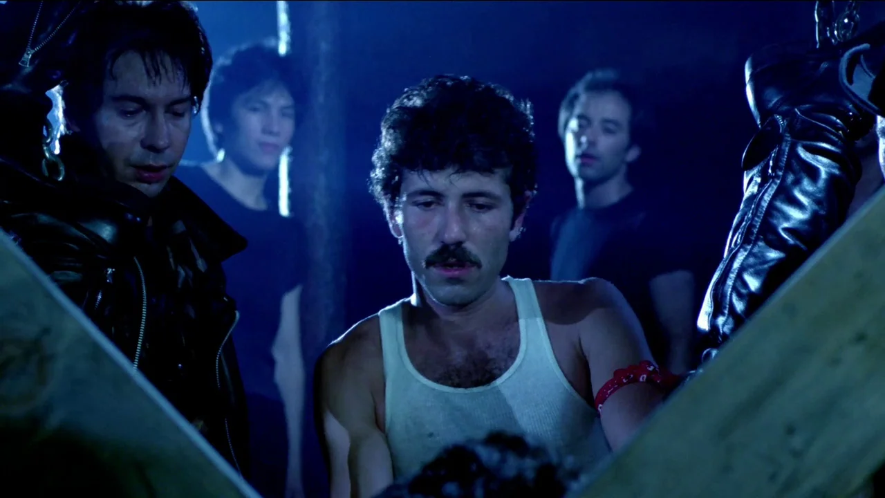 Cruising (1980) movie still: medium — A dark, blue-lit room contains four men standing close together. The men wear black shirts and leather gear…