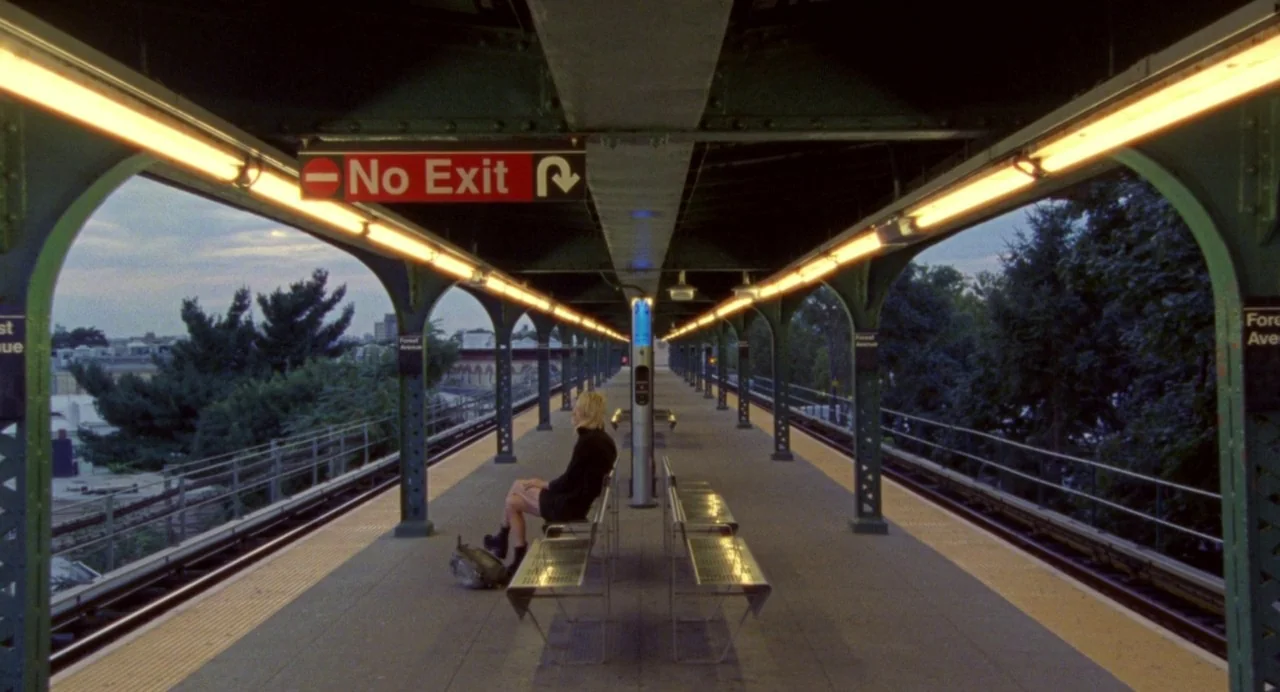 So Pretty (2019) movie still: wide — An elevated train station platform features rows of metal benches and green metal arches. A lone person in a…