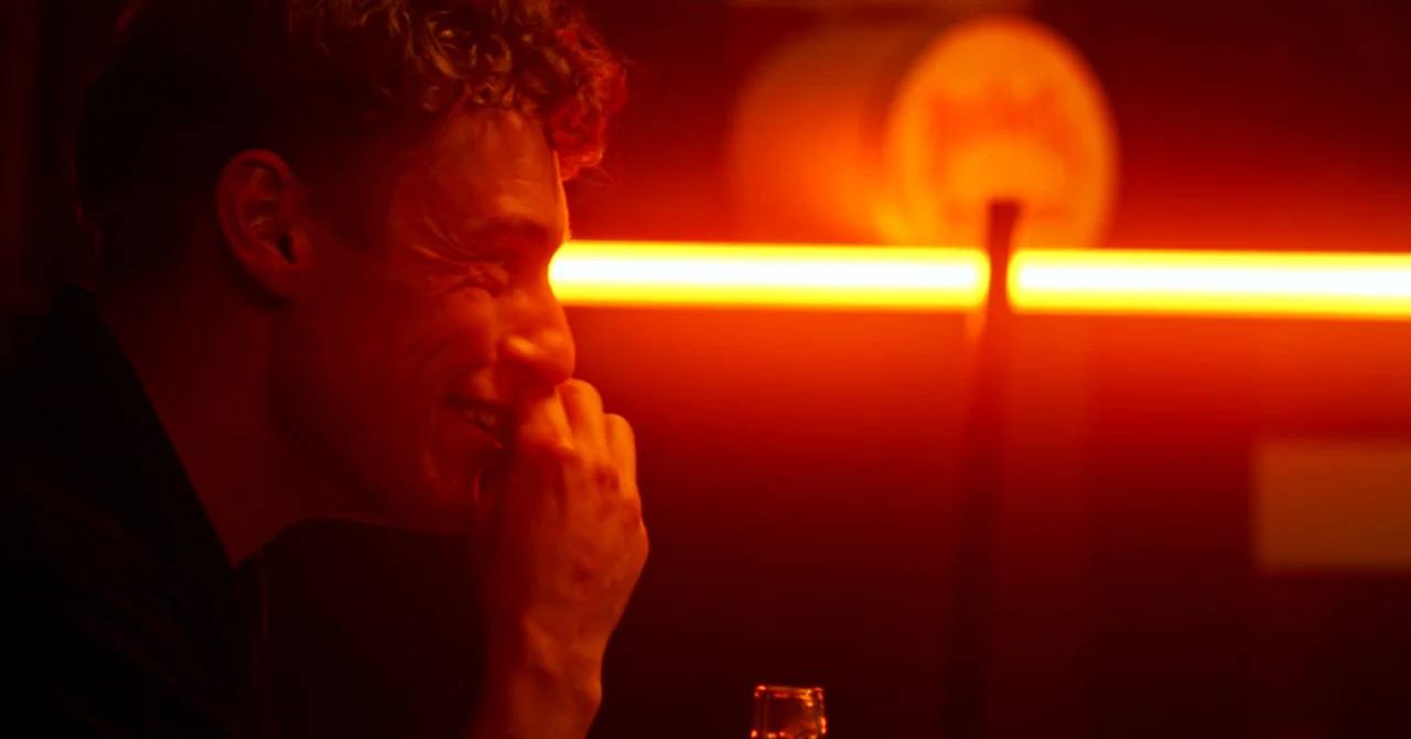 T Blockers (2023) movie still: closeup — A dimly lit bar interior features a glowing orange horizontal neon light in the background. A young man…