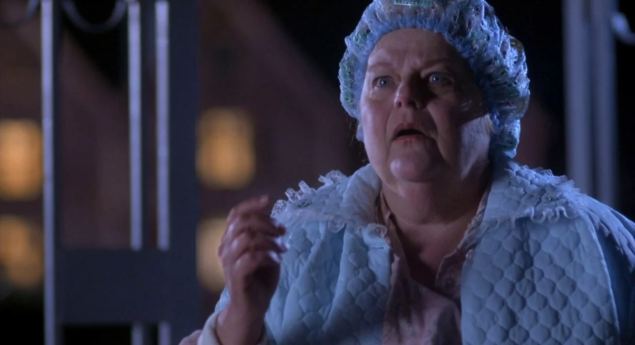 From Beyond (1986) movie still: closeup — An older woman stands on a porch at night wearing a blue housecoat. She looks off-camera with a surprised…