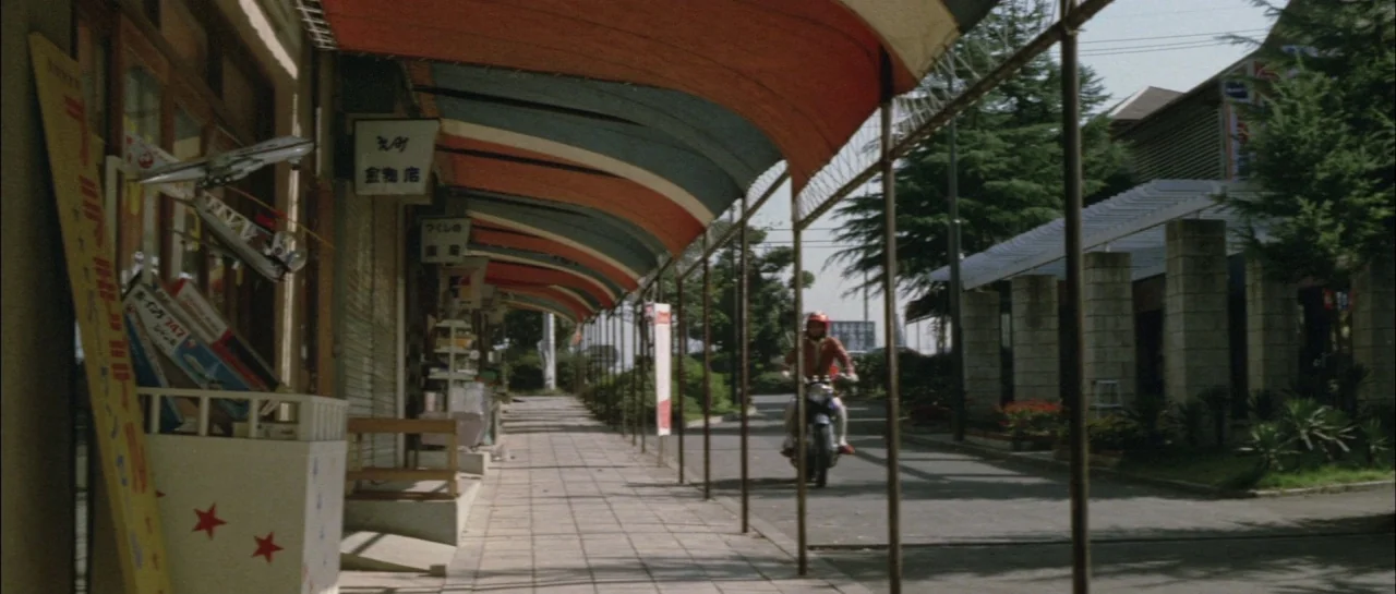 Godzilla Vs Megalon (1973) movie still: wide — A Japanese outdoor shopping walkway features a red and white canopy over tiled flooring. A person on…