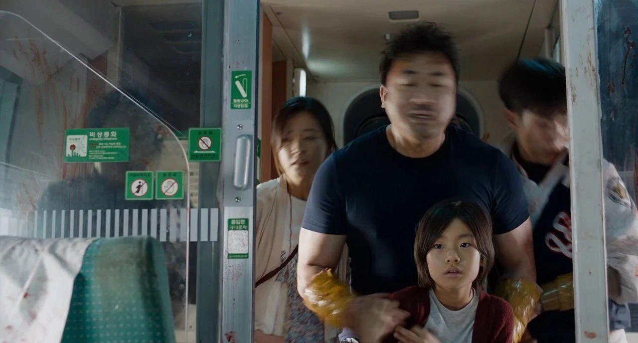 Train To Busan movie still: medium — Inside a modern commuter train car with blood-smeared glass partitions and emergency signs. A muscular man…