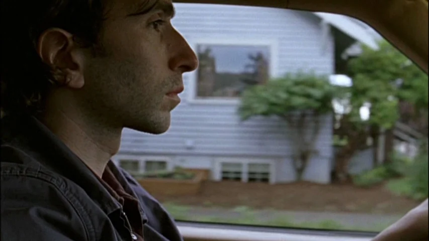 Old Joy (2006) movie still: medium — A man drives a car past a suburban house with light blue siding. The exterior of a residential home is visible…