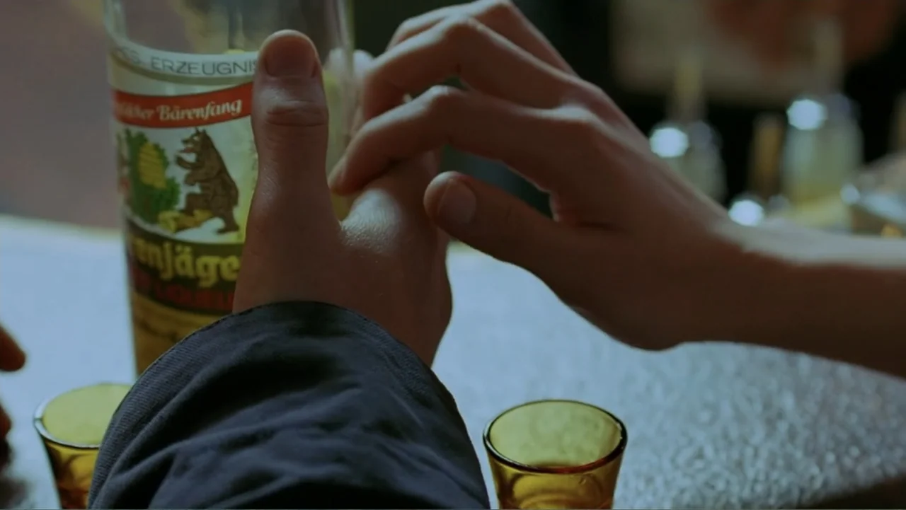 Emily (1976) movie still: closeup — A bar counter surface features a liqueur bottle and two small empty shot glasses. Two human hands are positioned…