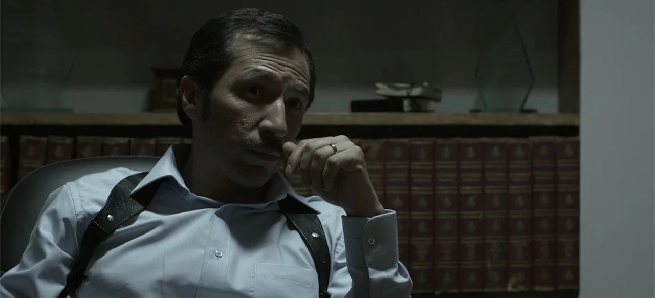 About the Deaths (2016) movie still: medium — A dimly lit office features shelves filled with dark leather-bound books in the background. A man…