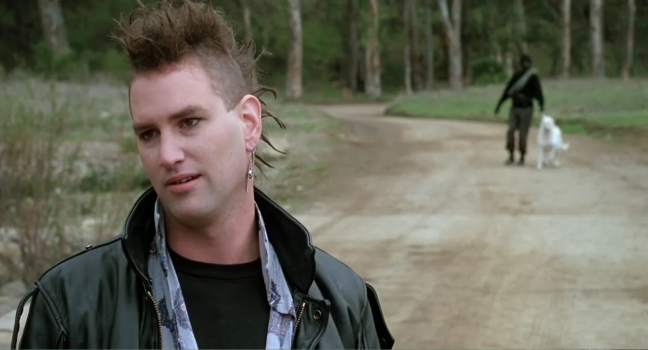 Dangerously Close (1986) movie still: medium — A rural dirt road passes through a wooded area with mature trees. A man with a mohawk stands in the…