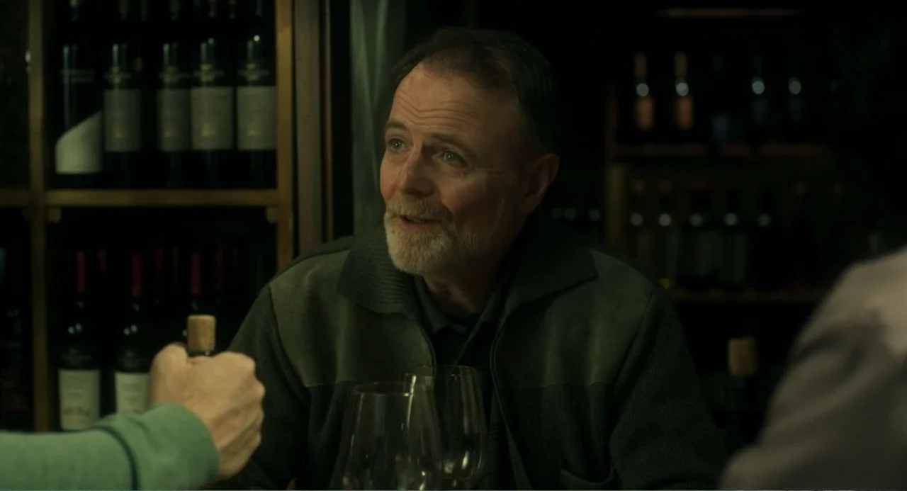 Message in a Bottle (1999) movie still: medium — A dimly lit room features shelves filled with wine bottles in the background. A man sits at a table…