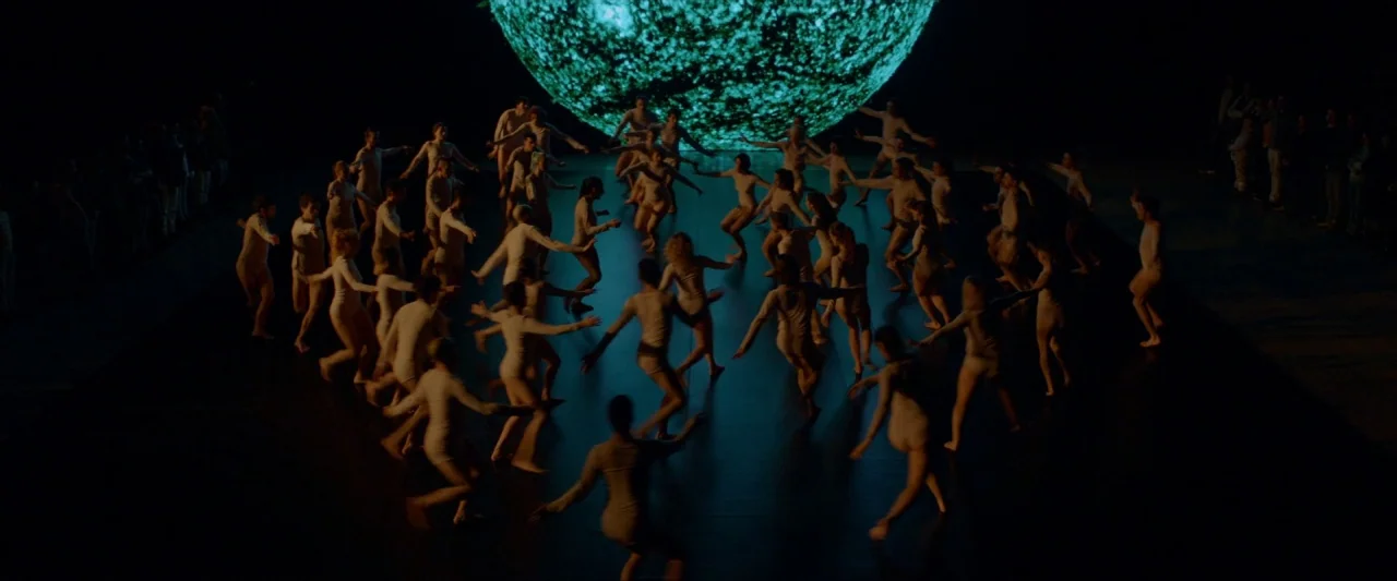Ema (2019) movie still: wide — A dark, expansive stage features dozens of dancers in tight-fitting pale clothing. They form a large circular…