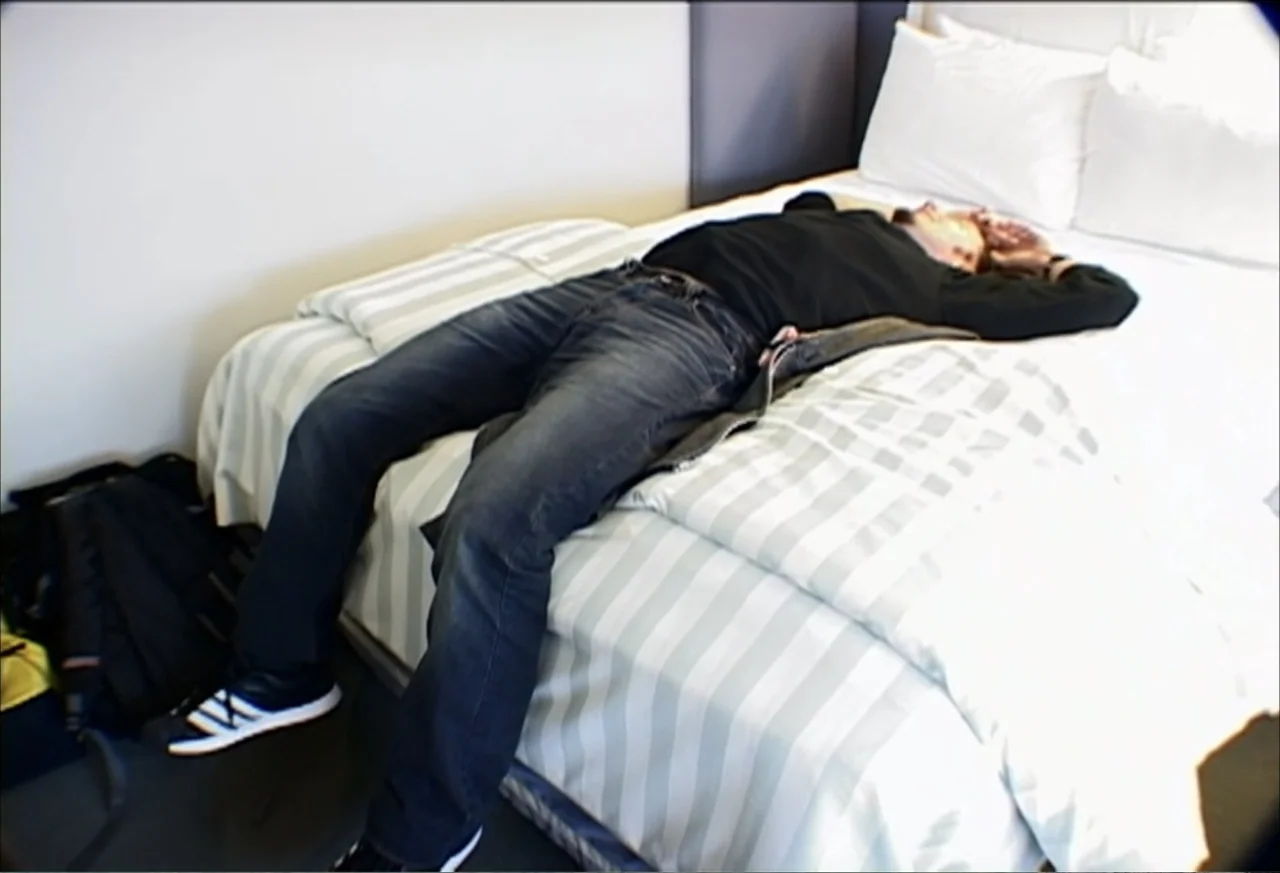 Plastic Disasters (2006) movie still: fullbody — A modern hotel room features a white bed with gray striped bedding. A man in black clothing lies…