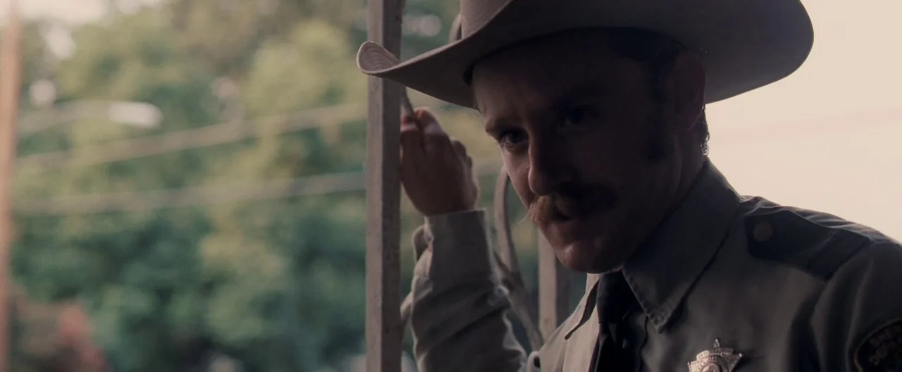 Ain’t Them Bodies Saints (2013) movie still: closeup — A man in a sheriff uniform stands on a porch near metal railings. Outside the structure,…