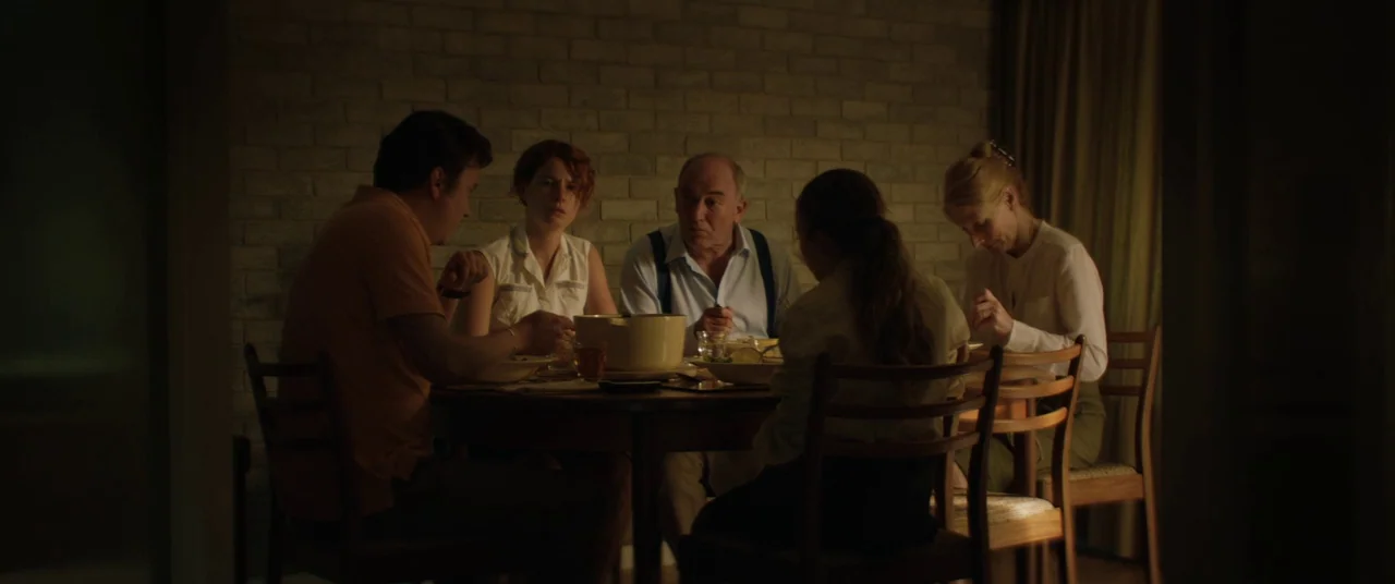 Beast (2017) movie still: medium — A group sits around a round wooden table in a dimly lit room. They are eating dinner in front of a textured grey…